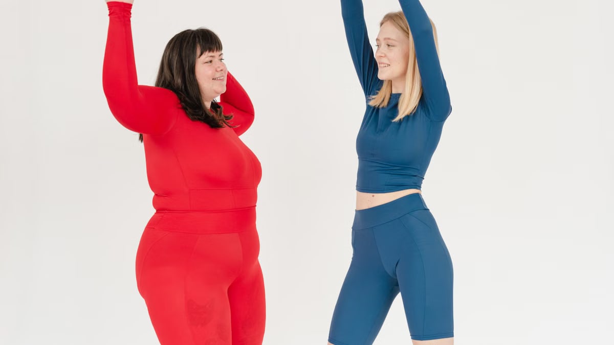 Energetic plus size female and slender woman in activewear dancing with raised arms while looking at each other on white background